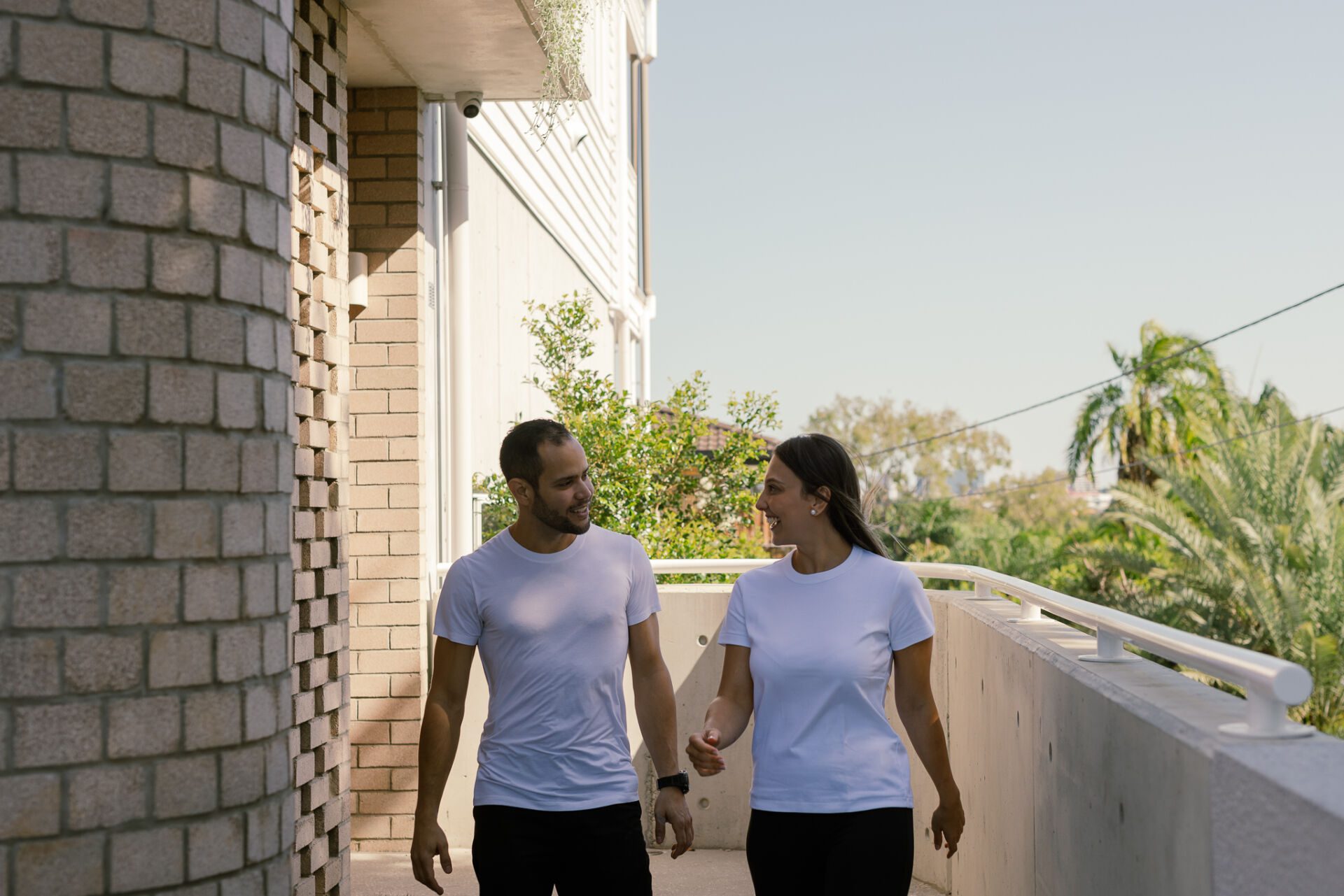Two people in white shirts walk along a sunlit brick building alley, smiling and conversing.