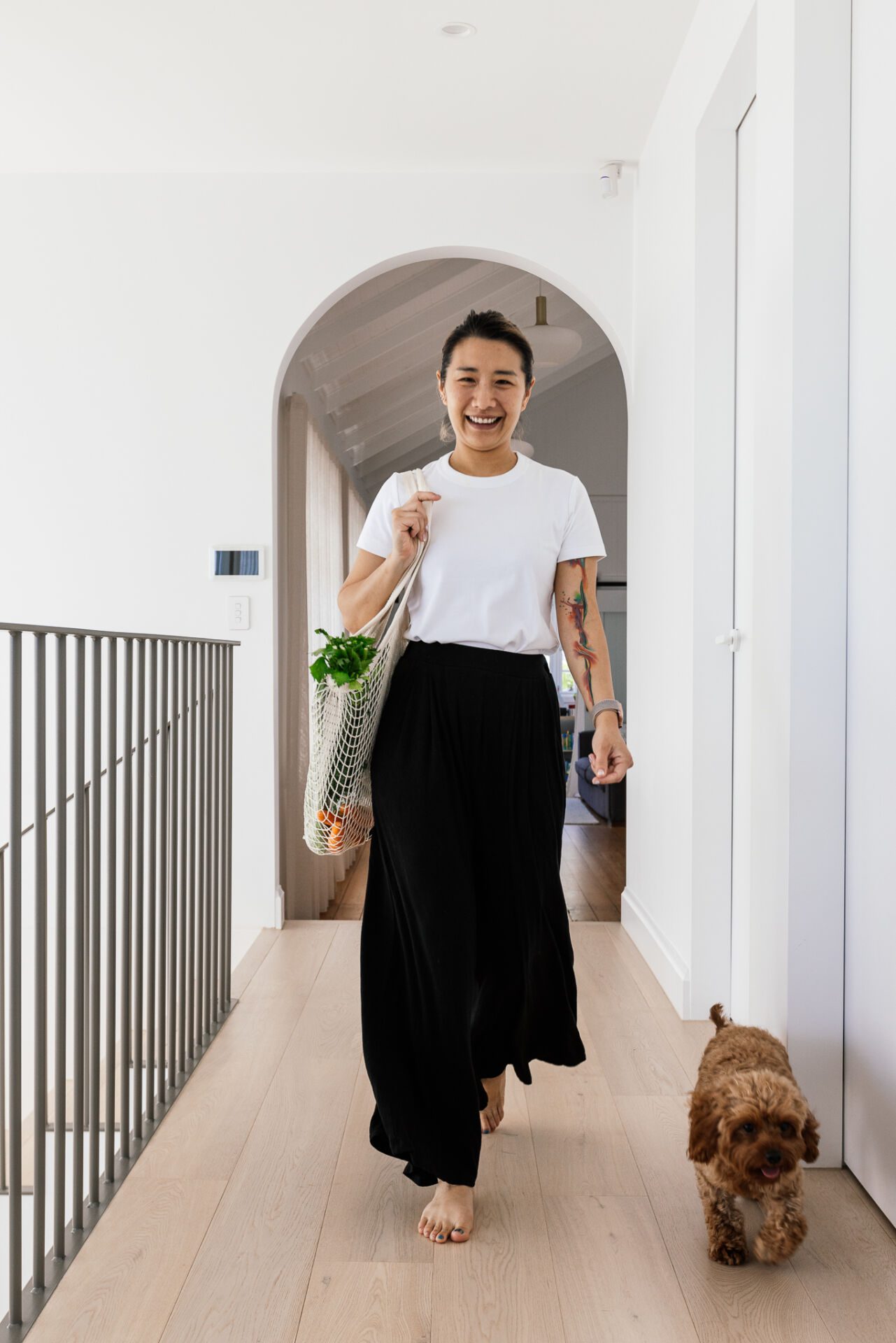 Smiling woman walking barefoot down a bright hallway, carrying a mesh tote with vegetables and a small brown dog walking beside her.