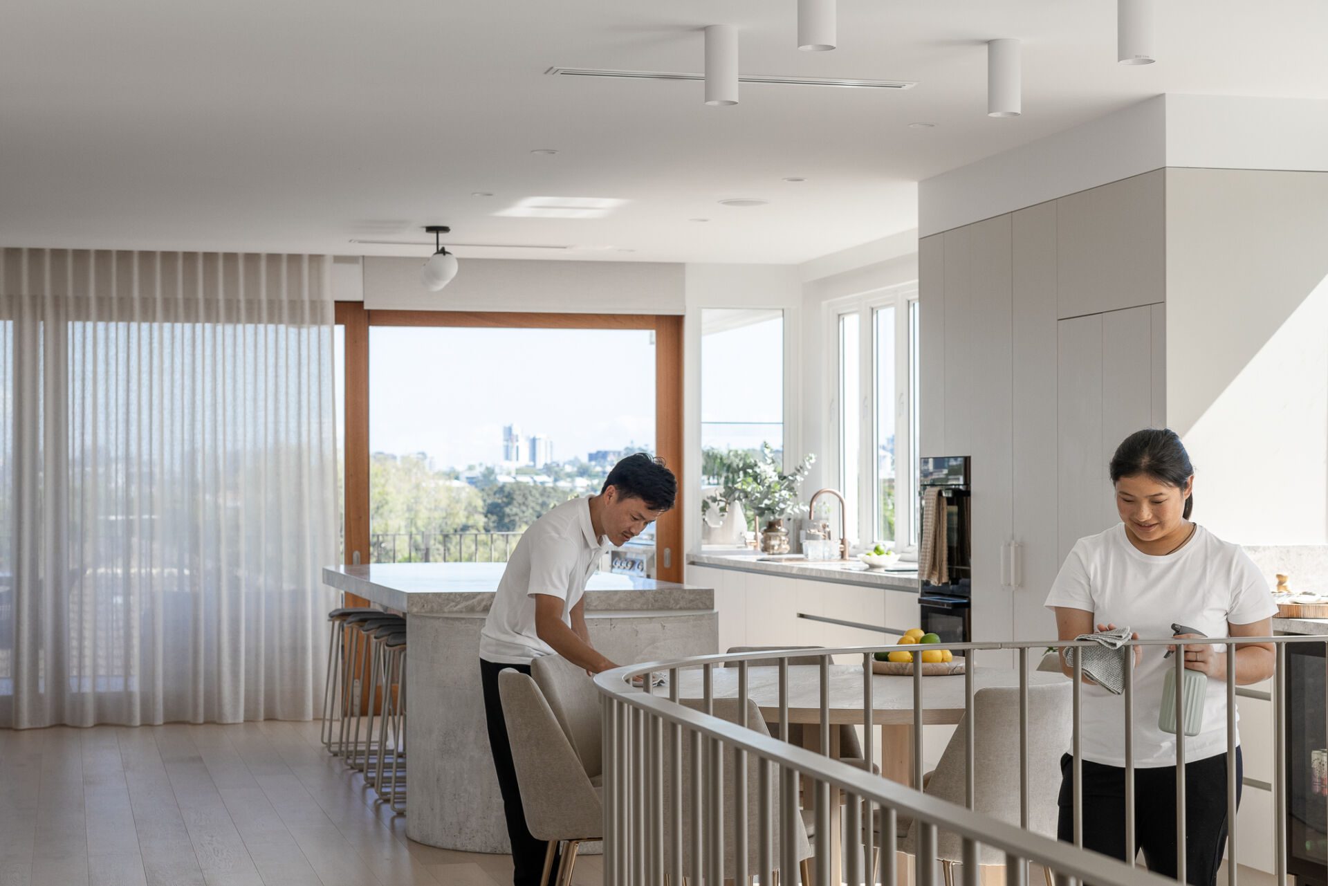 Two Family Clean Cleaners in Canberra in a bright modern kitchen at an island, with a fruit bowl and large windows in the background, one wiping the counter and the other folding a towel near a railing.