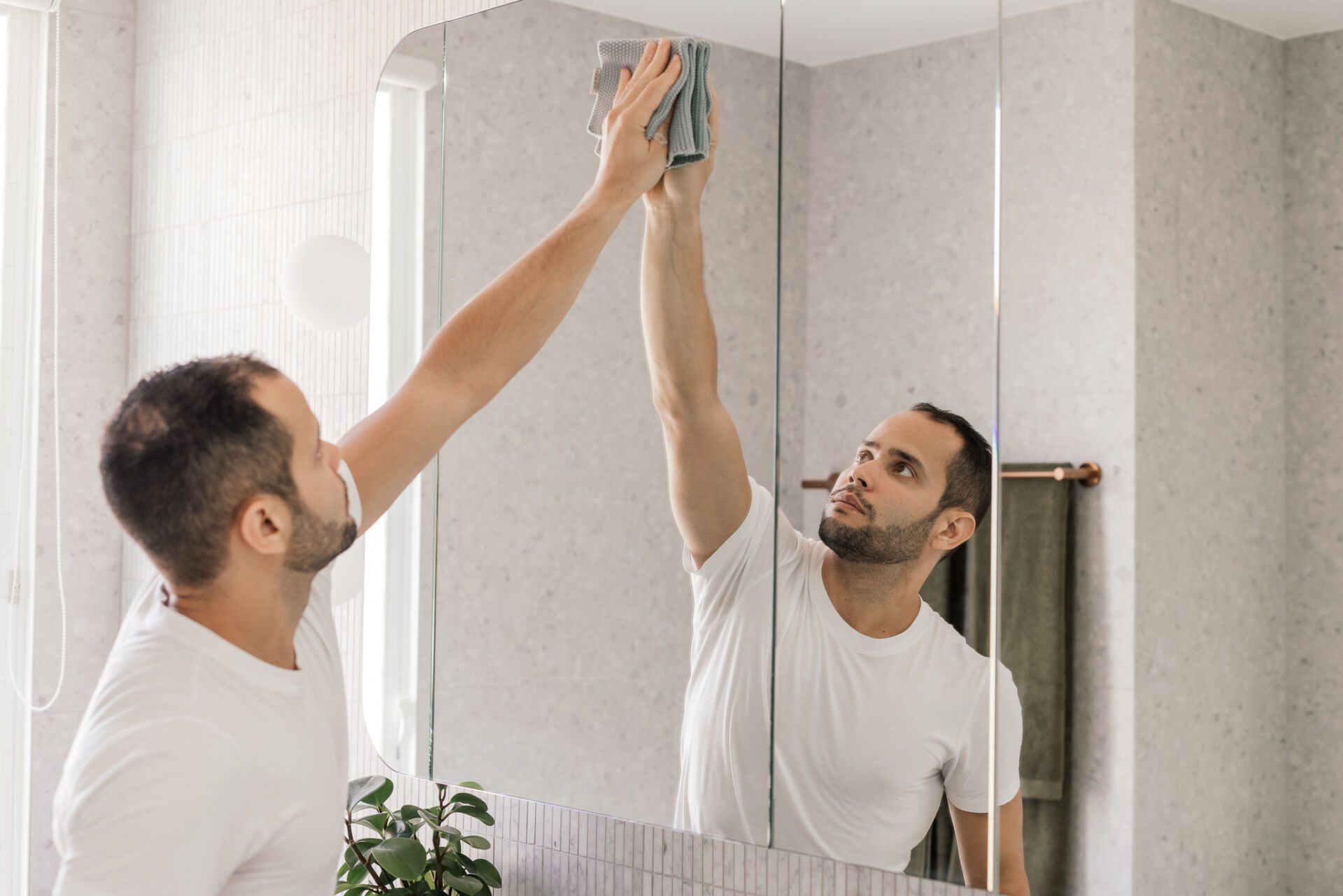 Man cleaning a bathroom mirror in an Adelaide home with a gray cloth while looking at his reflection in a tiled bathroom.