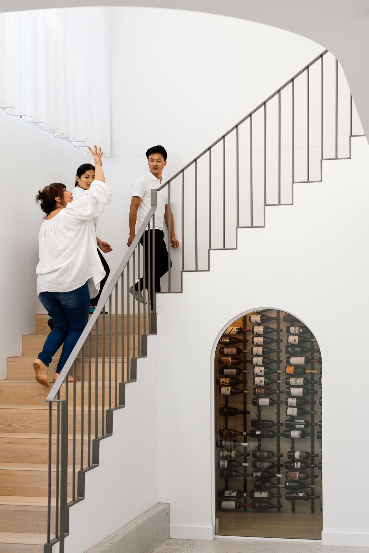Three people ascend a modern white staircase beside a circular wine cooler built into the wall.