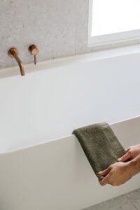Copper bathtub faucet and control knob mounted on a gray wall above a white tub, with a person folding a gray towel nearby.