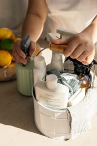 A white bucket filled with cleaning supplies, spray bottles, cloths, and a wooden-handled tool, on a kitchen counter with fruit in the background (blurred).